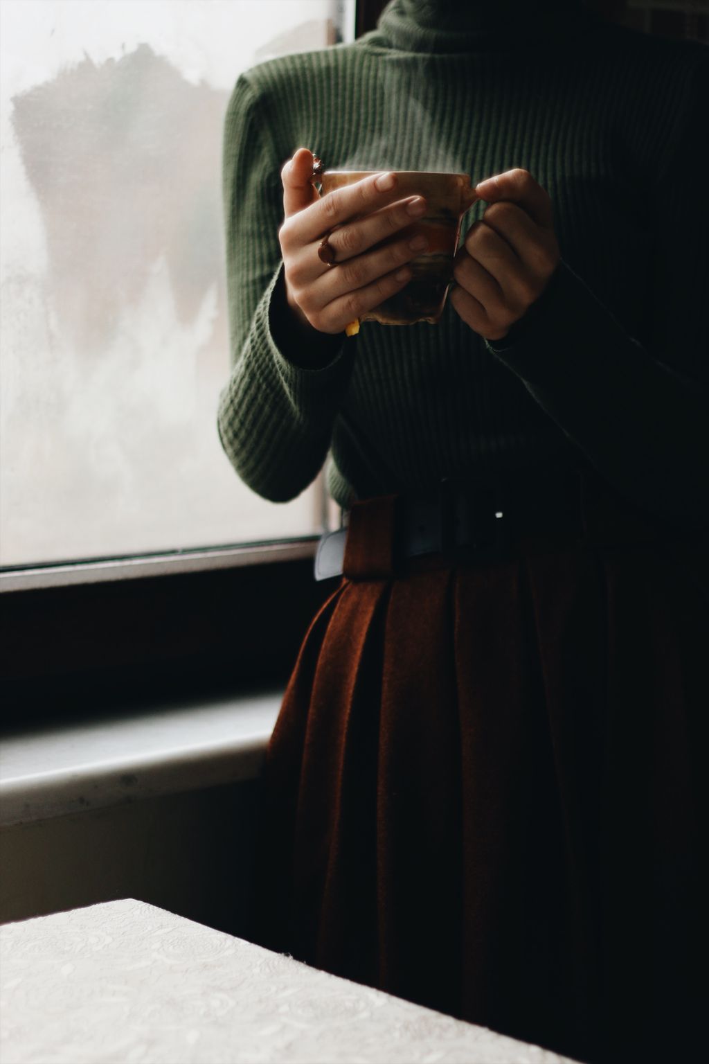 A person in a green turtleneck cradles a marbled mug of coffee beside a fogged-up window on a grey, quiet morning.