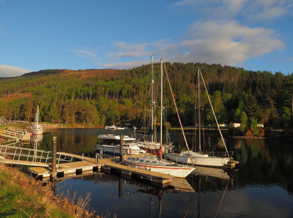 A quiet marina at sunrise, sailboats resting on still water with soft hills behind and warm light low on the horizon.